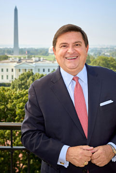 Stewart McLaurin standing on a balcony that overlooks the White House and the Washington Monument. He is wearing a dark colored suit, reddish-pink tie, and blue oxford cloth shirt. 