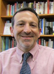 Headshot photo of Joshua Rothman in front of a bookcase. He is wearing a mauve shirt and tie.