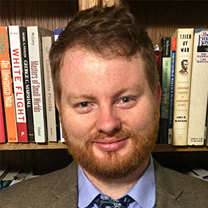 Jon Merritt with read hair and beard, wearing a blazer and tie, standing in front of a bookshelf.