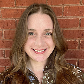 Shoulders-up profile photo of a person with long wavy hair wearing a patterned brown blouse, photographed against a red brick wall background.