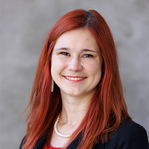 Professional headshot of a person with long red hair, facing the camera and smiling. The person is wearing a dark blazer over a red top, a pearl necklace, and a single visible rectangular earring. The background is a softly blurred gray studio backdrop, with even lighting and the subject centered in the frame.